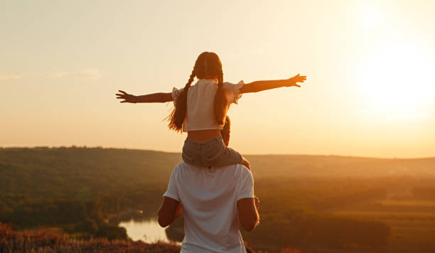 Unrecognizable father and daughter during evening in nature Back view of anonymous man carrying girl with outstretched arms wand admiring sundown while spending time in countryside together on shoulders stock pictures, royalty-free photos & images