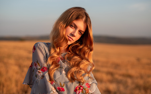 Pretty young female in trendy floral outfit looking at camera while standing in dry meadow on summer day in nature