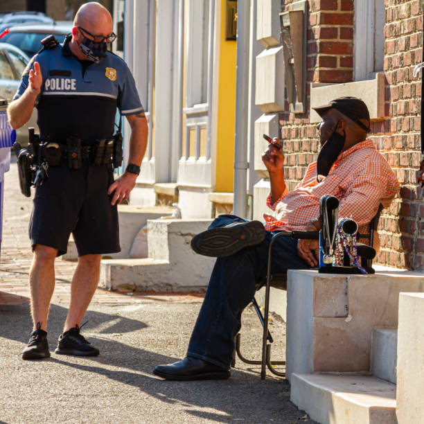 A white police officer with shaved head is talking to a black man smoking cigar Ellicott City, MD, USA 10/07/2020: A white police officer with shaved head who is wearing  short and t shirt type uniform and face mask is  talking to an elderly black man who is smoking cigar outside ellicott city maryland stock pictures, royalty-free photos & images