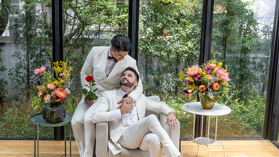 Young gay couple in white tuxedo holding each other during wedding ceremony at home. Two gays sharing their happy feeling at a flower corner in a house with love and care
