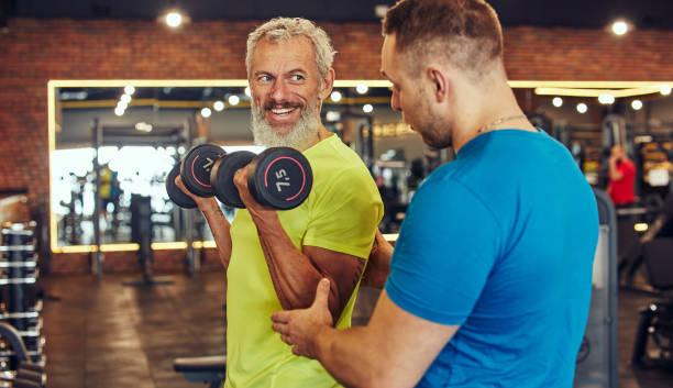 Working out at gym with fitness instructor. Positive mature man in sportswear doing weight exercises with assistance of young professional personal trainer Working out at gym with fitness instructor. Positive mature man in sportswear doing weight exercises with assistance of young professional personal trainer. Sport, healthy and active lifestyle fitness trainer stock pictures, royalty-free photos & images