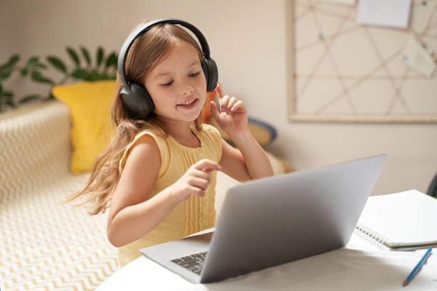 cute little caucasian girl in headphones using laptop while sitting at the table in her room, watching video lesson at home - child computer laptop little girls imagens e fotografias de stock