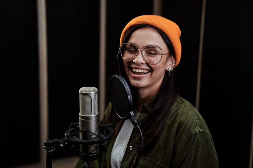 Portrait of cheerful young woman, hip hop artist singing into a condenser microphone while recording a song in a professional studio. Selective focus. Horizontal shot