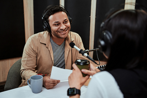 Portrait of young male radio host going live on air, talking with female guest, holding a script paper while sitting in studio. Selective focus. Horizontal shot