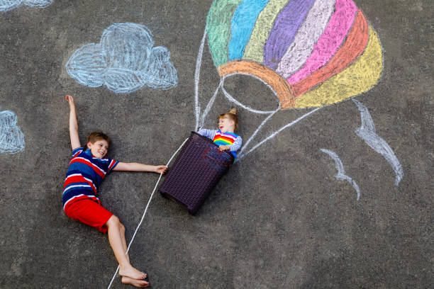 petite fille heureuse d’enfant et garçon d’enfant volant dans la montgolfière peinte avec des craies colorées dans des couleurs d’arc-en-ciel sur le sol ou l’asphalte en été. deux enfants, frères et sœurs s’amusant. loisirs créatifs - hot air balloon flying heat people photos et images de collection