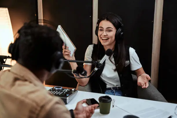 Portait of happy female radio host laughing, listening to male guest, presenter and holding a script paper while moderating a live show in studio. Focus on woman. Horizontal shot