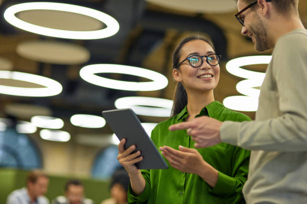 Discussing project. Young positive asian woman showing something on digital tablet to her male colleague while standing together in the coworking space or office stock photo