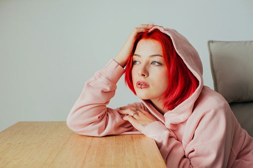 Portrait of a cool teen girl with bright red hair, lip piercing and hoodie holding head on desk looking at camera