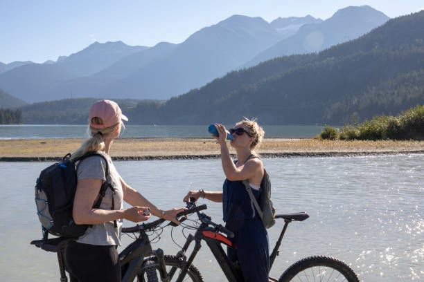 mujeres ciclistas electrónicos de montaña se rehidratan a orillas del lago por la mañana - rehydrate fotografías e imágenes de stock