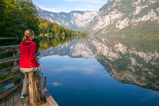 Morning view of Lake Bohinj with senior woman sitting on pier and enjoying  mountain reflection in lake water. It is in Julian Alps, Slovenia and is destination is occupied by Slovenia tourists because the coronavirus travel restrictions.  It is starting point to many hiking tours.