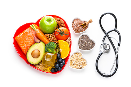 Healthy eating: fresh multicolored foods to help lower cholesterol levels and for heart care shot on white background. The composition includes oily fish like salmon. Beans like Pinto beans, soybeans and brown lentils. Vegetables like garlic, carrot, avocado, broccoli and tomato. Fruits like apple, orange and berries. Nuts like almonds. Olive oil. The food is arranged in a red heart shape tray and three bowls with chia seeds, flax seeds and oat flakes are beside it. A stethoscope complete the composition. High resolution 42Mp studio digital capture taken with SONY A7rII and Zeiss Batis 40mm F2.0 CF lens