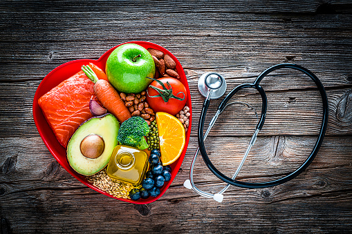 Healthy eating: fresh multicolored foods to help lower cholesterol levels and for heart care shot on wooden table. The composition includes oily fish like salmon. Beans like Pinto beans, soybeans and brown lentils. Vegetables like garlic, carrot, avocado, broccoli and tomato. Fruits like apple, orange and berries. Nuts like almonds. Olive oil. The food is arranged in a red heart shape tray and a stethoscope is beside it. High resolution 42Mp studio digital capture taken with SONY A7rII and Zeiss Batis 40mm F2.0 CF lens