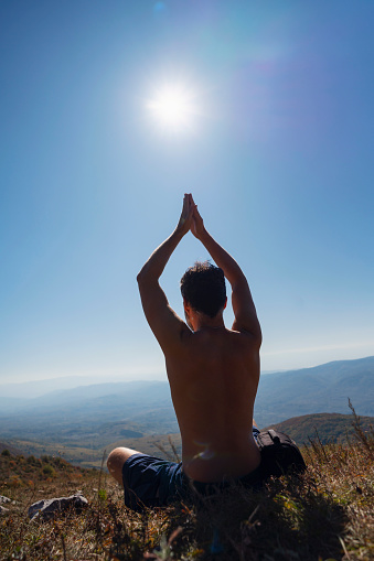 Mature Caucasian man sitting in a lotus position in nature, holding hands in mantra gesture, and meditating. Practicing breathing exercises on a sunny day.