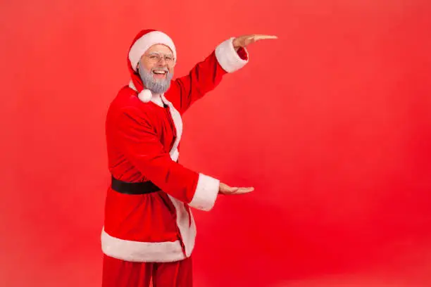 Portrait of smiling positive man pretending to be santa claus showing advertising area and looking at camera with smile, freespace for your advertisement. Indoor studio shot isolated on red background