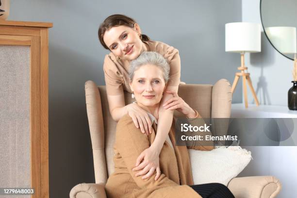 Elderly Middle Mother Sitting In A Chair And Her Daughter Are Hugging Looking At Camera Trusted Relations Stock Photo - Download Image Now