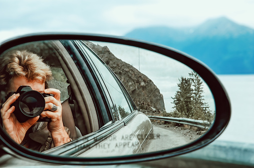 Caucasian woman photographing through the side view mirror of the car, with the text : Objects in mirror are closer than they appear.
