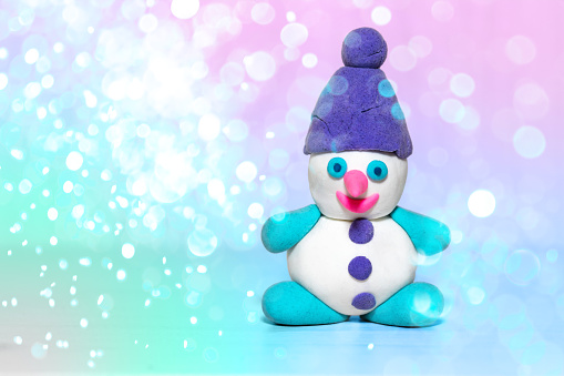 white plasticine snowman with purple winter hat, pink nose, playdough or modeling compound dough, concept of festive atmosphere of holiday, blurred christmas neon background, blue bokeh lights