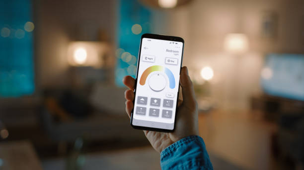 istock Young Handsome Man Gives a Voice Command to a Smart Home Application on His Smartphone To Set a Comfortable Light Temperature. It's a Cozy Evening in Technologically Advanced Living Room. 1279660348