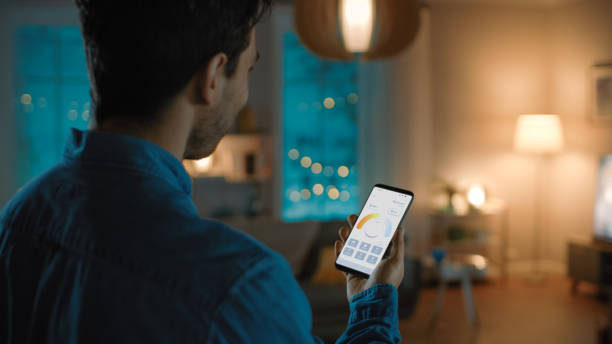 istock Young Handsome Man is Setting up Comfortable Bedroom Conditions with Voice Command to Smart Home Application on His Smartphone. It's a Cozy Evening in Technologically Advanced Living Room. 1279660361