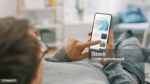Young Man At Home Is Lying On A Coach And Using Smartphone For Scrolling And Reading News About Technological Breakthroughs Hes Sitting On A Couch In His Cozy Living Room Over The Shoulder Shot Stock Photo - Download Image Now