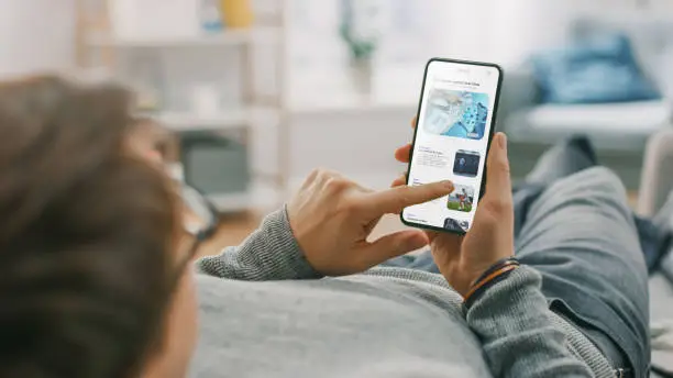 Photo of Young Man at Home is Lying on a Coach and Using Smartphone for Scrolling and Reading News about Technological Breakthroughs. He's Sitting On a Couch in His Cozy Living Room. Over the Shoulder Shot