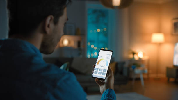 young handsome man gives a voice command to a smart home application on his smartphone to change light temperature and set a comfortable lighting. it's a cozy evening in living room. - warm lighting imagens e fotografias de stock
