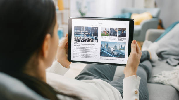 Over the Shoulder Shot of a Young Woman at Home Lying on a Couch and Using Tablet Computer in Horizontal Landscape Mode for Reading News about Technology. Girl Using Touchscreen Device. Over the Shoulder Shot of a Young Woman at Home Lying on a Couch and Using Tablet Computer in Horizontal Landscape Mode for Reading News about Technology. Girl Using Touchscreen Device. surfing the net stock pictures, royalty-free photos & images