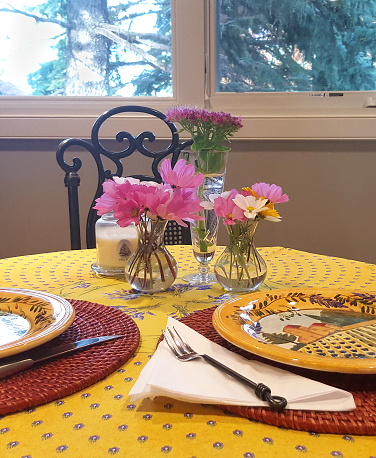 Place setting for two . Table with small vases of pink and white flowers. Yellow table cloth and plates. Window in background