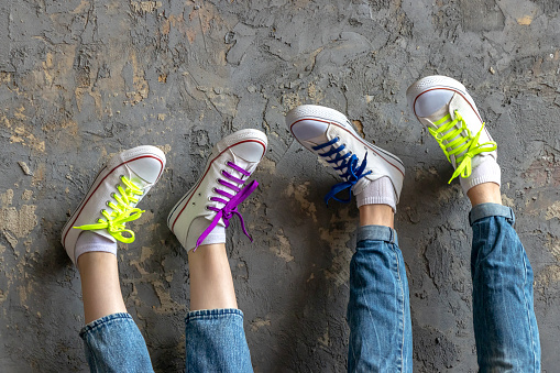two pairs of legs of a girl and a young man wearing blue jeans and white sneakers, feet up on a vintage textured background wall, shoelaces of green, pink and blue color