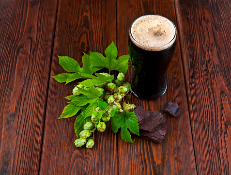 Glass of dark stout beer with natural green hop leaves and chopped chocolate on a wooden table.
