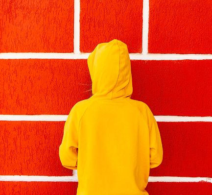Teenager girl in a yellow sweatshirt or hoodie in a hood against the background of a red wall with white seams