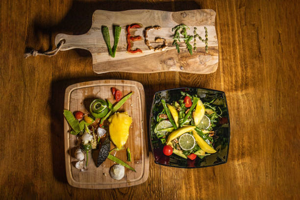 Vegan word written with vegetables on cutting board with salad bowl on wooden table, and  peeled leftovers