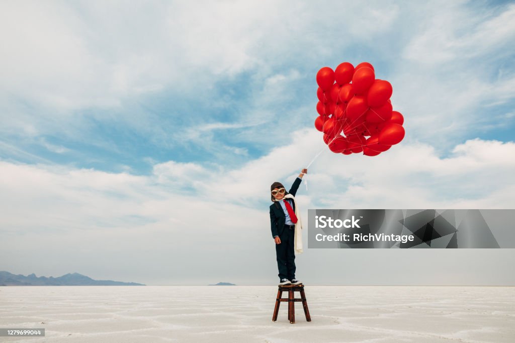 Young Business Boy with Balloons A young boy dressed in business suit stands on a stool wearing a flight cap and flying goggles holds dozens of red balloons looking to launch his business into the profits atmosphere. Image taken at the Bonneville Salt Flats in Utah, USA. 6-7 Years Stock Photo
