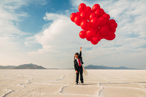 A young boy dressed in business suit, flight cap, and flying goggles holds dozens of red balloons looking to launch his business into the profits atmosphere. Image taken at the Bonneville Salt Flats in Utah, USA.