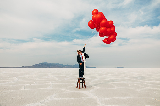 A young boy dressed in business suit stands on a stool wearing a flight cap and flying goggles holds dozens of red balloons looking to launch his business into the profits atmosphere. Image taken at the Bonneville Salt Flats in Utah, USA.