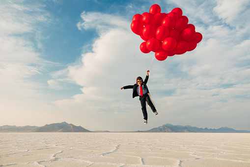 A young boy dressed in business suit, flight cap, and flying goggles holds dozens of red balloons flies his business into the profits atmosphere. Image taken at the Bonneville Salt Flats in Utah, USA.