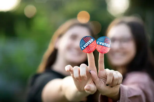 Portrait of two women  holding out their hands together with the I voted stickers attached to their fingers for the viewer to see. Their fingers form the v for victory and peace sign.