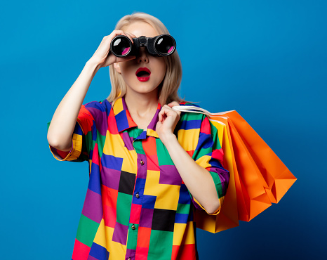 Blonde girl in 90s shirt  with binoculars and shopping bags on blue background