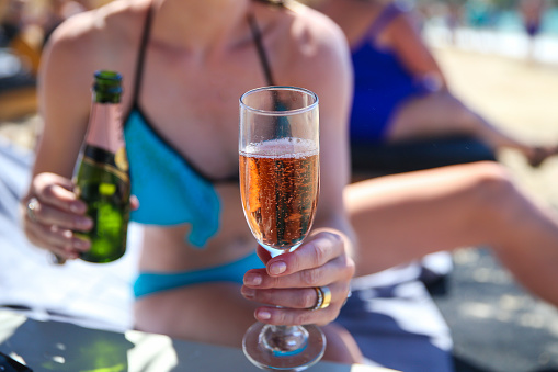 A woman handing a flute of sparkling rose to the viewer in a beach in Greece