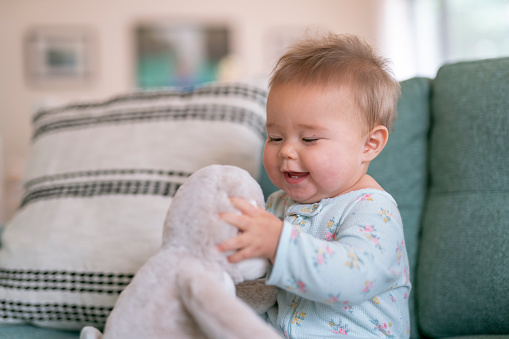 An adorable mixed race baby sits up by herself and laughs while playing with a stuffed toy. The child is sitting on a couch at home.