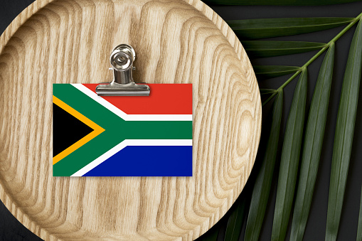 South Africa flag tagged on wooden plate. Tropical palm leaves monstera on background. Minimal national concept.