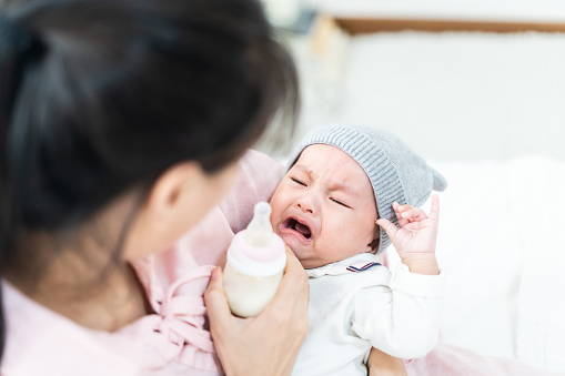 Close up view of beautiful Asian mother and her crying lovely newborn baby. Mother holding her baby and feeding cute newborn baby from milk bottle. Happy family concept.