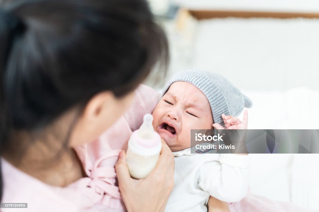Close up view of beautiful Asian mother and her crying lovely newborn baby. Mother holding her baby and feeding cute newborn baby from milk bottle. Happy family concept. Baby - Human Age Stock Photo