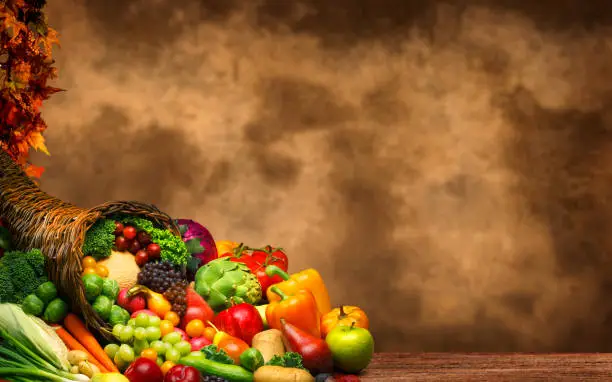 A Thanksgiving still life of a cornucopia full of holiday fruits and vegetables in front of a rustic background that provides ample room for copy and text.