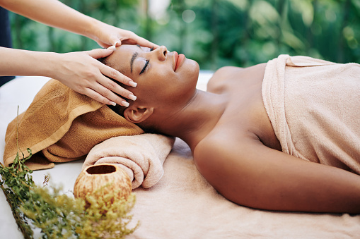 Happy relaxed young Black woman enjoying head and face massage in beauty salon