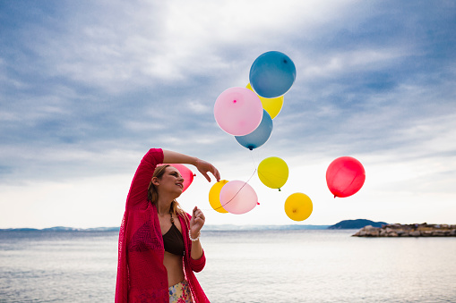 Cheerful and playful young woman walking at the beach with multi-colored balloons and enjoying her summer vacation in Greece