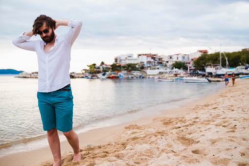Young bearded man wearing a white shirt and blue shorts walking at the beach and enjoying a sunny summer day during his vacation in Greece