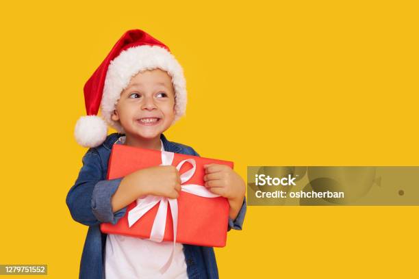 Portrait Of A Satisfied Little Child Boy In Christmas Santa Hat Laughing Isolated Over Yellow Background Holds A Gift Box Preparing For The New Year Holidays Stock Photo - Download Image Now