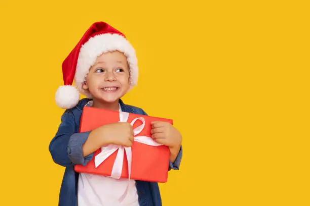 Photo of Portrait of a satisfied little child boy in christmas Santa hat. laughing isolated over yellow background. Holds a gift box. Preparing for the New Year holidays