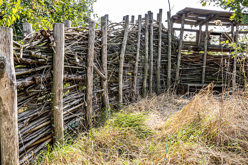 Rustic fence made with tree branches and wooden posts with a small roof with small openings, brown green wild grass, sunny summer day in a nature reserve in South Limburg, the Netherlands